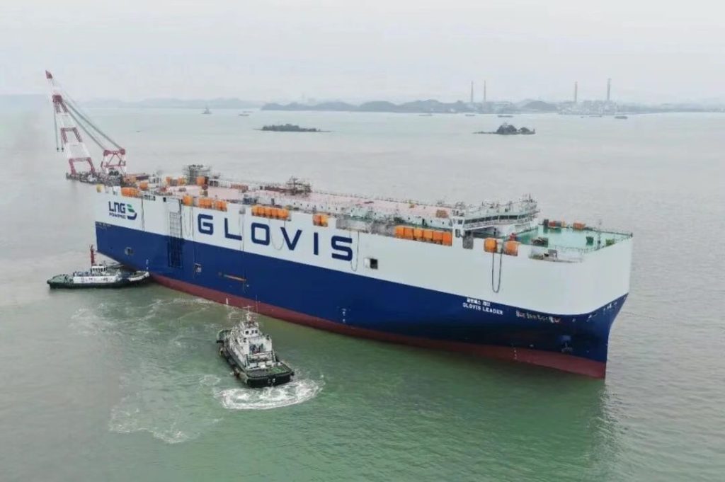 Barco de carga GLVIS en el puerto, rodeado de remolcadores y con un cielo nublado de fondo.